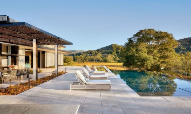 A contemporary single-storey residence beside a reflective swimming pool, with broad pale paved terraces, sculptural sun loungers, extensive outdoor living, and a deep timber pergola supported by slender dark posts. Floor-to-ceiling glazing opens the house to a landscaped rural setting with mature trees and low hills in warm late-afternoon light.