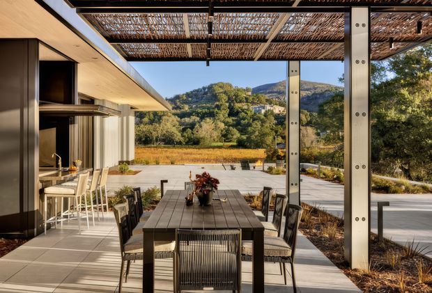A contemporary outdoor dining terrace viewed from beneath a pergola, with a long dining table and chairs in the foreground. An outdoor kitchen or bar with stools sits to the left beneath a broad roof overhang. Slim dark metal structural posts, a woven or slatted pergola roof, large open glazing, pale paved surfaces, and landscaped planting frame views toward a wooded hillside.