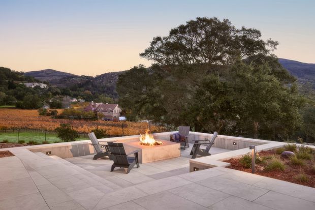 A spacious contemporary outdoor terrace overlooking a vineyard valley and wooded hills. The pale paved patio features built-in low seating walls, several dark lounge chairs, and a central square outdoor fire pit with flames. Integrated linear lighting and restrained landscaping create a refined evening entertaining area.