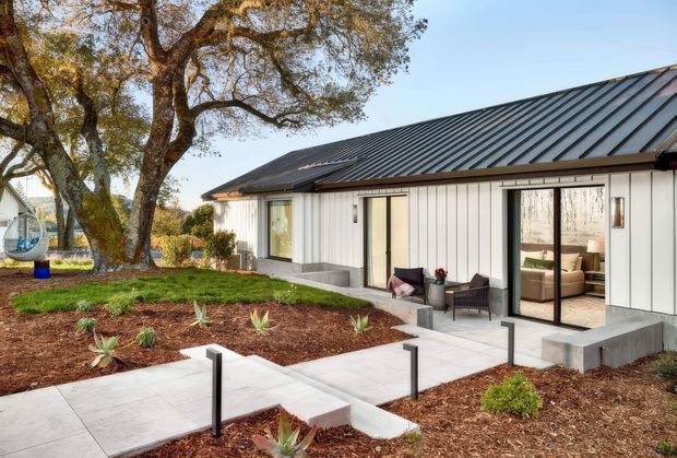 Single-storey contemporary pavilion-style home viewed from the landscaped exterior. The building has vertical light-coloured cladding, a dark standing-seam metal pitched roof, large glazed sliding doors, and a concrete patio with simple black post lighting. Mulched garden beds and mature trees surround the house, with a glimpse of a furnished interior through the glazing.