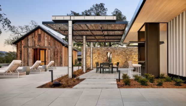 Contemporary outdoor courtyard entertaining area linking a timber-clad barn-like structure with a modern pavilion. The space features a large paved terrace, steel-framed pergola with a slatted roof, outdoor dining furniture, lounge chairs, a textured natural-stone boundary wall, and a modern building with warm timber soffits and vertical light-coloured cladding. The setting is landscaped with low planting and mature trees.