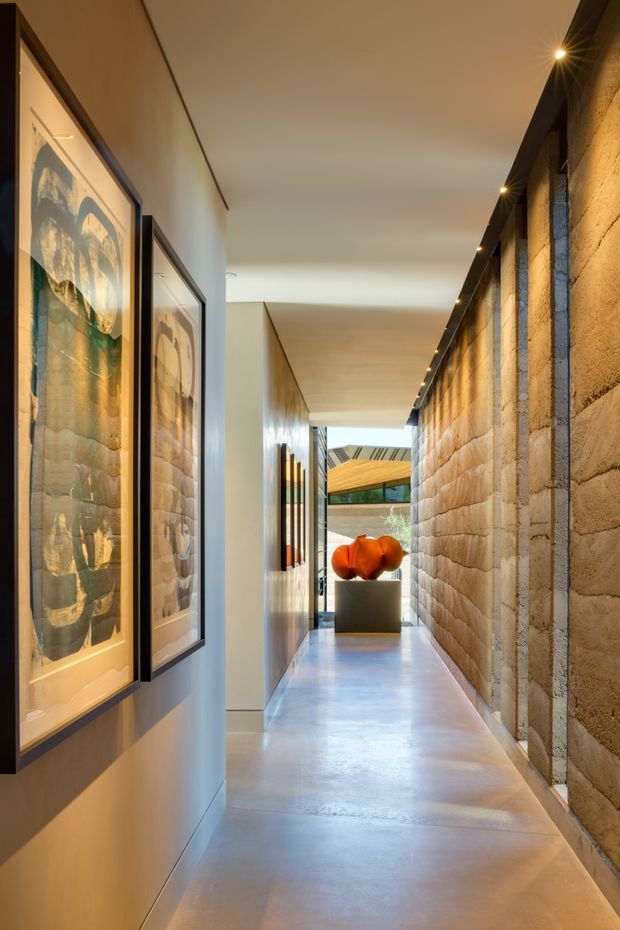 A long, narrow architectural hallway with a polished concrete floor and warm recessed ceiling lighting. One side features exposed textured concrete block masonry, while the opposite smooth pale wall displays large framed artworks. The corridor leads toward a brighter courtyard or glazed opening, with a sculptural orange object on a pedestal at the far end.