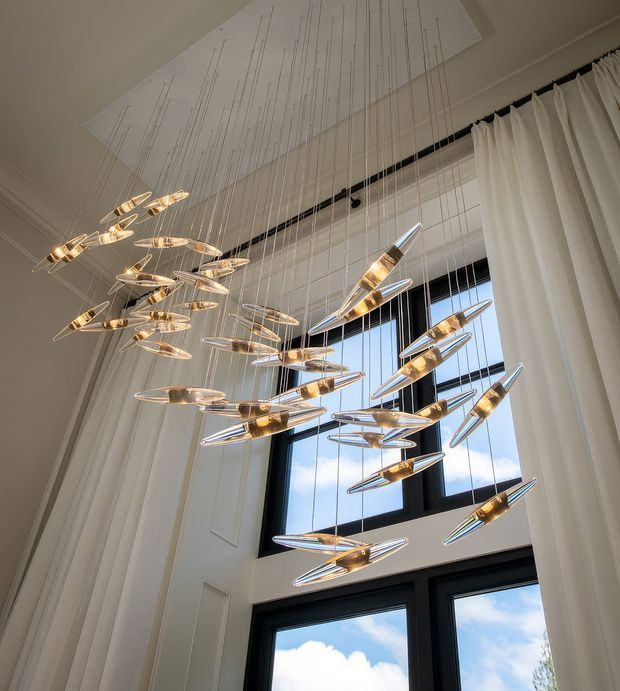 Interior view of a tall, refined space featuring a dramatic suspended cluster chandelier composed of numerous elongated metallic and translucent pendant elements. The fixture hangs on many fine cables beneath a large rectangular ceiling panel, in front of expansive black-framed windows with full-height white curtains. White paneled walls and abundant daylight create an elegant, airy atmosphere.