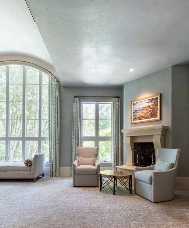 A refined living room with a tall curved bay window and large central window, both dressed in patterned curtains. The room features muted grey-green walls, pale carpet, upholstered armchairs, a small round coffee table, and a classic stone or painted fireplace with a framed landscape artwork above it. Recessed ceiling lights and restrained furnishings create a calm, traditional atmosphere.