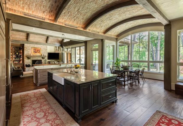 A spacious, traditionally styled open-plan kitchen and dining area with a large dark-stained island, white stone benchtop, integrated sink, and pendant lighting. The room features rich timber flooring, extensive built-in cabinetry, exposed curved brick barrel-vault ceilings with dark timber beams, and a large arched window wall overlooking greenery. A dining table sits beside the glazing, while a secondary sitting or living area with a fireplace is visible beyond the kitchen.
