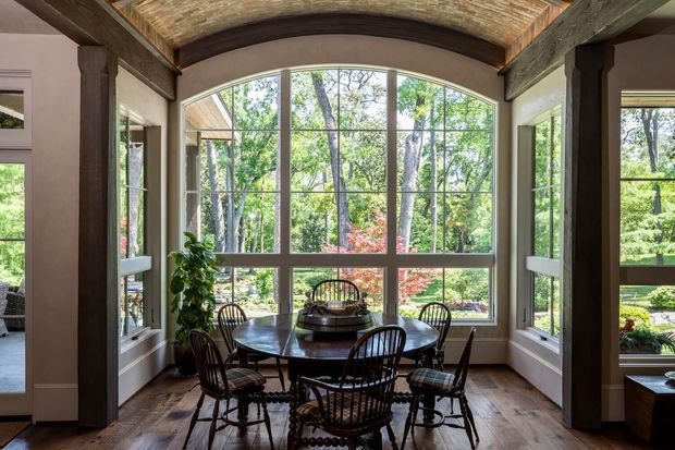 A bright enclosed dining nook surrounded by large gridded windows, with a round dark wood dining table and spindle-back chairs. The room features a dramatic curved timber barrel-vault ceiling with exposed dark beams, warm timber flooring, and substantial dark timber framing around the openings. Lush garden vegetation is visible outside through the glazing.
