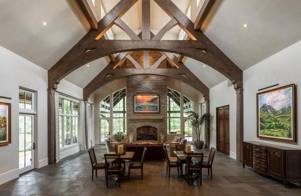 A spacious, formal living room with a dramatic vaulted ceiling and exposed dark timber trusses. The room features a large stone fireplace with timber surround, tall multi-pane windows and glazed doors, a dark wood dining or seating arrangement, traditional cabinetry, framed artwork, and large-format stone or slate flooring. The symmetrical composition and abundant glazing create a refined, warm, lodge-inspired interior.