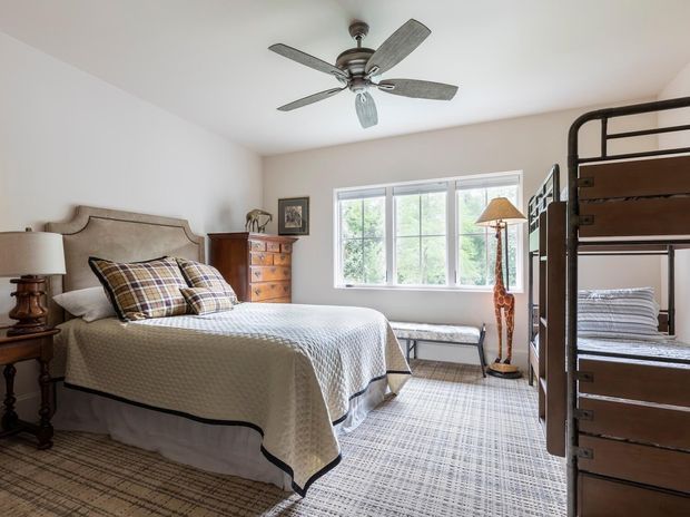 Bright traditional bedroom with a queen-size bed, upholstered decorative headboard, patterned plaid cushions, bedside tables and lamps, a wooden chest of drawers, large multi-pane window, ceiling fan, and a dark wood bunk bed along the right side. The room has pale walls, wall-to-wall patterned carpet, and a warm, comfortable residential character.