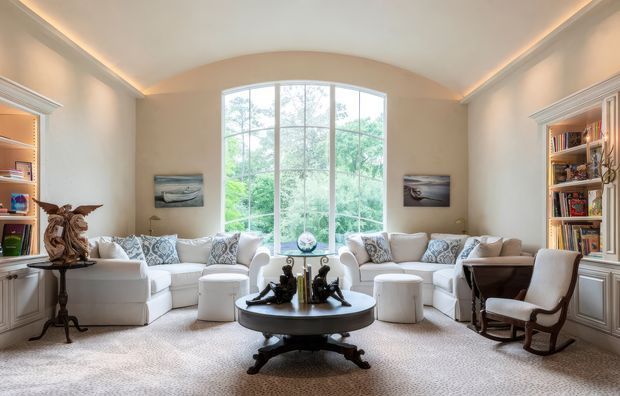 A bright, elegant living room arranged around a large arched-top floor-to-ceiling window. The room features pale upholstered sofas and ottomans, a dark oval coffee table, built-in bookcases on both sides, a rocking chair, decorative art, and warm concealed ceiling lighting. The interior has a refined, symmetrical, traditional character with neutral tones and views of greenery outside.