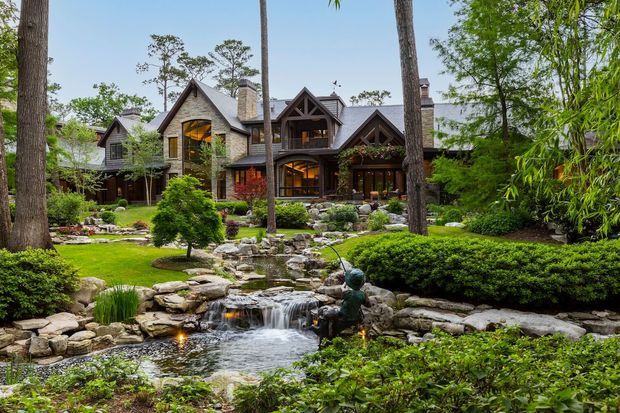 Exterior view of a large rustic lodge-style residence set within a meticulously landscaped garden. The home features multiple steep gables, stone-clad walls, dark timber framing and trim, prominent chimneys, and warm lighting visible through expansive windows. In the foreground is a landscaped pond and cascading rock waterfall surrounded by mature trees, shrubs, lawn, and ornamental planting.