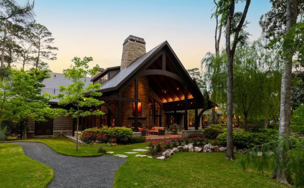 Exterior view of a substantial lodge-style home set within a landscaped garden. The dark timber-clad structure has a steep gabled roof, prominent covered entry or outdoor living porch, exposed timber framing, warm interior lighting, and a stone chimney. A gravel path, stepping stones, planted beds, mature trees, and a small landscaped water feature surround the building.