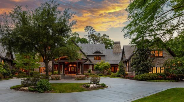 A large, warmly lit lodge-style residence viewed from a broad curved driveway at dusk. The complex has multiple connected gabled wings, steep shingle-clad roofs, substantial stone chimneys, dark timber detailing, covered entry porches, and extensive mature landscaping. Glowing windows and the dramatic sunset create a welcoming, rustic atmosphere.