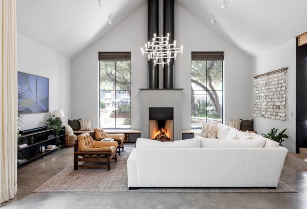 Bright, spacious living room with a dramatic vaulted ceiling, central fireplace with twin black flues, large windows, and a sculptural chandelier. A large white sofa faces the fireplace, with caramel leather lounge chairs, a television and media console to the side, a neutral area rug, and a textured wall hanging. The room has a refined contemporary feel with warm timber and leather accents.