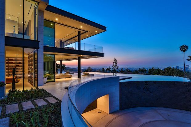 Twilight exterior view of a luxurious multi-level contemporary residence with expansive floor-to-ceiling glazing, projecting flat roof planes, open balconies with frameless glass balustrades, and warm recessed lighting. A curved rendered wall and lower-level illuminated entry or garage zone frame the foreground, while an infinity-edge pool or terrace overlooks a broad urban skyline and distant horizon.