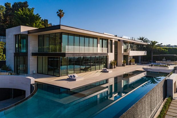 A large contemporary flat-roofed residence viewed from outside, with extensive floor-to-ceiling glazing, pale stone or concrete facade surfaces, dark window frames, broad terraces, and a striking reflecting swimming pool. Palm trees and landscaped grounds surround the property.