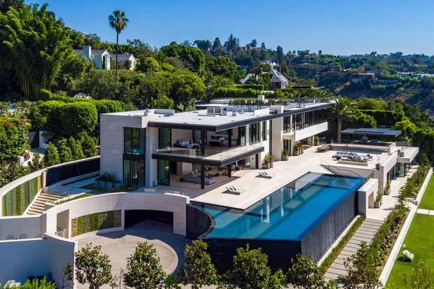 Aerial view of a large contemporary multi-level residence set into a lush, elevated suburban site. The home features a flat roof, expansive floor-to-ceiling glazing, projecting terraces, pale rendered or stone-like walls, extensive outdoor entertaining areas, and a long rectangular swimming pool with integrated stepped landscaping. Dense mature vegetation and neighbouring hillside homes surround the property.