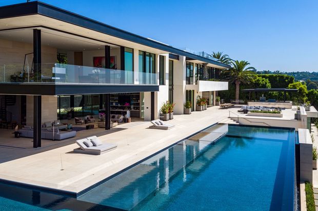 A large contemporary multi-storey residence viewed from the pool terrace, featuring a flat roof, extensive floor-to-ceiling glazing, slim dark frames, cantilevered upper balconies with clear glass balustrades, pale stone or concrete terrace paving, and a long rectangular swimming pool. The open-plan living areas are visible through the glazing, with landscaped surroundings and distant suburban greenery beyond.