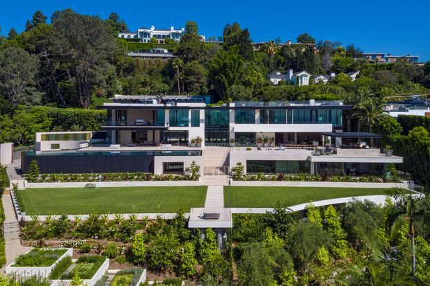 Aerial front view of a large contemporary multi-level residence set into a lush, landscaped hillside. The broad, low-slung house has extensive floor-to-ceiling glazing, flat rooflines, dark horizontal cladding, pale rendered walls, terraces, balconies, and a formal central stair descending through manicured lawns and gardens. Other hillside homes and mature vegetation form the background under a clear blue sky.