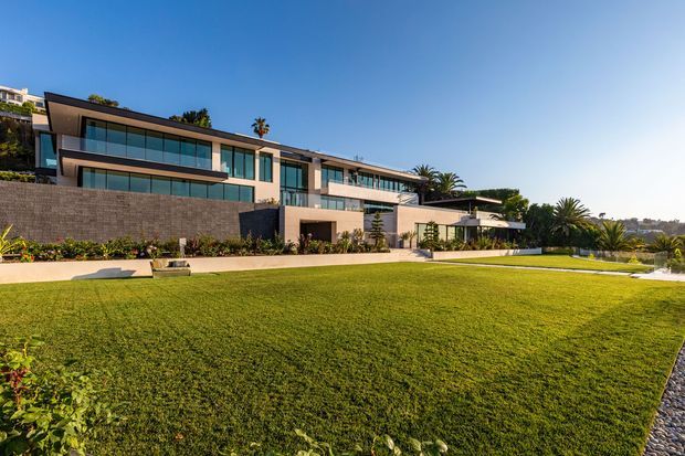 A large contemporary multi-level residence viewed from an expansive landscaped lawn. The house features broad floor-to-ceiling glazing, projecting upper volumes, extensive glass balustrades, pale rendered walls, dark masonry retaining walls, and flat rooflines. Palm trees and manicured planting surround the property, with a bright blue sky and distant urban landscape beyond.
