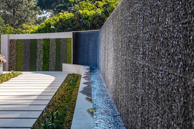 A landscaped courtyard water feature with a long textured stone or pebble-clad wall forming a cascading narrow channel. Pale rectangular paving leads through the garden, bordered by low planting and a patterned green-and-stone vertical garden wall. The scene is bright, contemporary, and carefully composed, with dense trees visible beyond the enclosure.