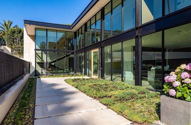 Exterior view of a contemporary multi-level residence with extensive floor-to-ceiling glazing, dark slim window frames, pale rendered wall surfaces, and a flat roof. A paved walkway runs through a landscaped garden toward the glass-lined building, with an internal stair and living spaces visible through the glazing.