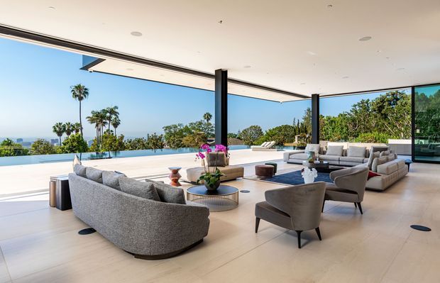 A spacious, high-end open-plan living room with floor-to-ceiling corner glazing that opens toward a terrace and expansive ocean or coastal views. The room features multiple light grey upholstered sofas and lounge chairs arranged around low round coffee tables, a pale smooth floor, slim black structural columns, recessed ceiling lighting, and a long linear ceiling vent. The interior has a refined contemporary appearance with strong indoor-outdoor connection and abundant natural light.