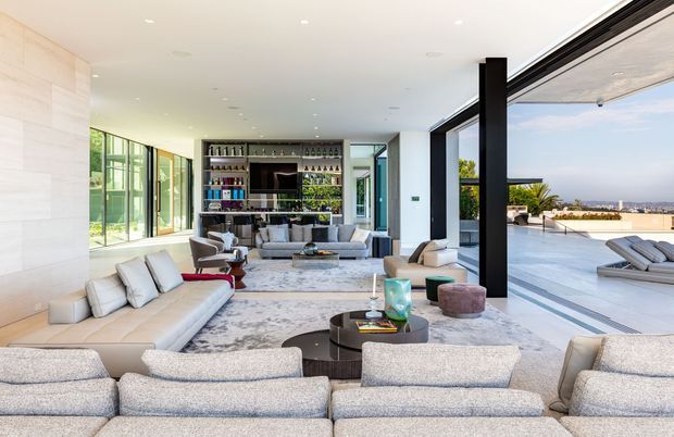 A bright, spacious contemporary living room with multiple pale upholstered sofas arranged around low round coffee tables and a large patterned rug. The room features floor-to-ceiling glazing, a dark built-in bar and display wall, recessed ceiling lighting, and wide open stacking doors leading to a terrace with a pool and distant landscape views.