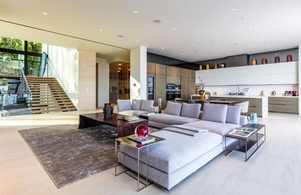 A bright, spacious open-plan living area with a large pale sectional sofa, glass-and-metal coffee tables, a dark area rug, and a sleek linear kitchen in the background. A floating timber staircase with glass balustrading leads to an upper level beside floor-to-ceiling glazing. The room has recessed lighting, pale flooring, light timber wall panels, white cabinetry, and a refined contemporary fit-out.