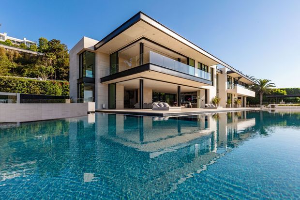 A contemporary multi-level luxury residence viewed from across a large swimming pool. The house features clean rectangular volumes, extensive floor-to-ceiling glazing, pale masonry or rendered walls, dark metal window and balcony frames, broad cantilevered roof and balcony overhangs, and transparent glass balustrades. Poolside outdoor seating is visible beneath the covered terrace, with landscaped greenery and neighbouring hillside residences in the background.