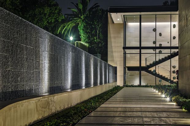 Night-time exterior approach to a contemporary multi-level building. A long textured dark masonry or stone feature wall is illuminated with vertical wall-washer lighting, leading along a broad paved walkway toward a glazed entry volume. Through the floor-to-ceiling glazing, a sculptural stair and indoor climbing wall are visible. Concrete-toned side walls, integrated step lighting, tropical planting and palm foliage contribute to a dramatic modern architectural setting.