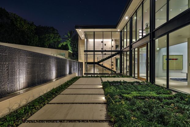 Night-time view of a contemporary multi-storey residence organized around a landscaped courtyard. A linear paved walkway with gravel joints leads between low clipped planting and a textured illuminated water feature wall toward extensive floor-to-ceiling glazing. The black-framed glass facade reveals a double-height interior with a sculptural stair, recessed lighting, and minimalist furnishings.