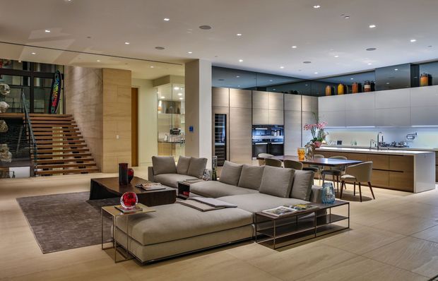 Open-plan contemporary interior combining a spacious living room and kitchen. A large modular sectional sofa and low glass-and-metal coffee tables occupy the foreground, while the rear features a streamlined kitchen with a long island, integrated cabinetry, pale stone-look surfaces, and bar seating. A timber slatted stair and sculptural display wall add architectural interest. Recessed ceiling lighting and a restrained neutral palette create a polished, upscale atmosphere.