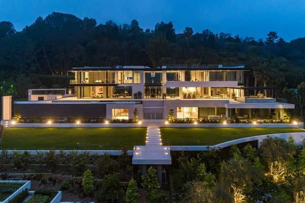 A large contemporary multi-level residence photographed at dusk, viewed from the landscaped front garden. The house has a long, low horizontal composition with flat rooflines, extensive illuminated glazing, projecting upper volumes, terraces, and a central entrance aligned with a straight path across a manicured lawn. Dense wooded hills form the background, while layered planting and subtle landscape lighting frame the foreground.
