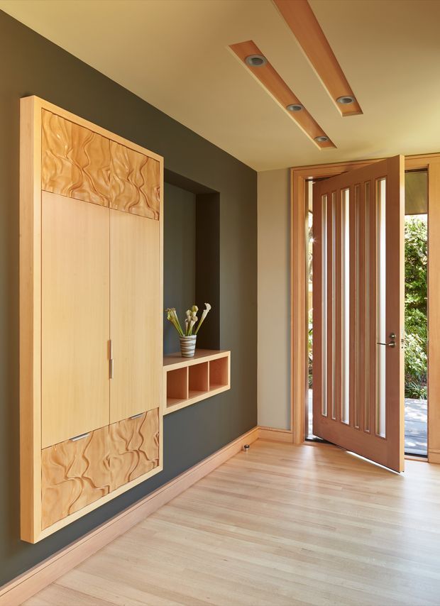 A warm, contemporary entryway with a built-in light timber storage cabinet featuring decorative carved wood panels, a recessed dark charcoal display niche, and a small floating shelf. The space has pale timber flooring, a timber-framed glazed front door with vertical inset detailing, and linear timber ceiling light recesses. Natural light enters through the open door and adjacent glazing.