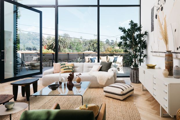 Bright contemporary living room with a large cream sectional sofa, layered neutral textiles, sculptural coffee tables, woven rug, indoor plants, and a white built-in sideboard. Full-height black-framed glazing and an open glazed door connect the room to a timber-decked outdoor terrace, with landscaped greenery visible beyond.