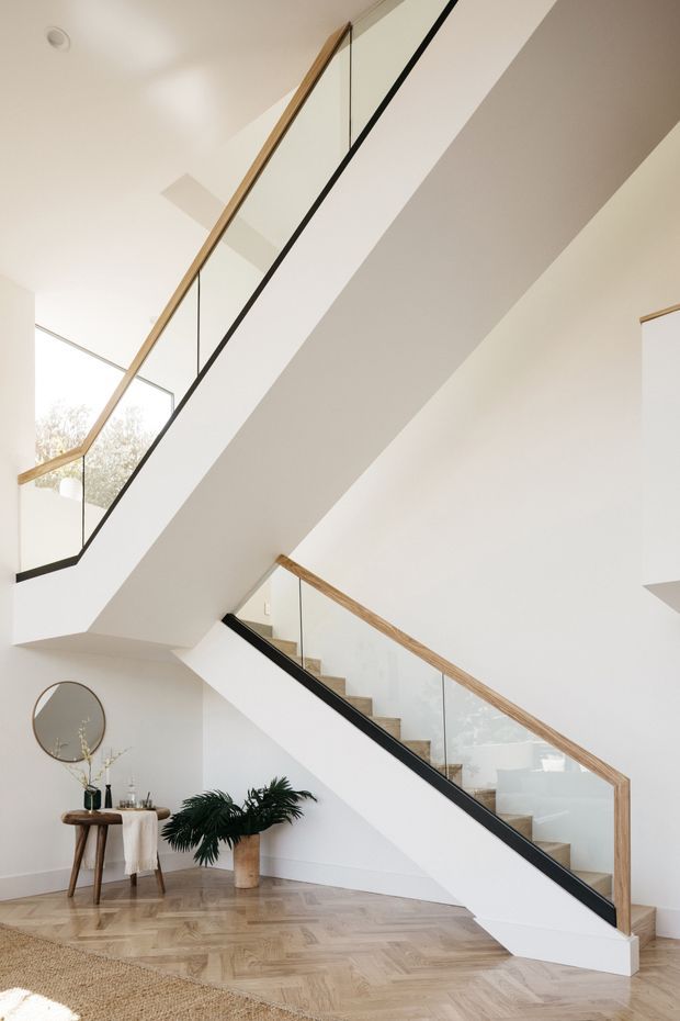 Bright contemporary interior featuring a sculptural staircase with white stringer walls, clear glass balustrades, slim black edging, and warm timber handrails. The staircase rises through a tall, light-filled space with white walls and ceilings, pale herringbone timber flooring, and a small styled console with mirror and plants beneath it.