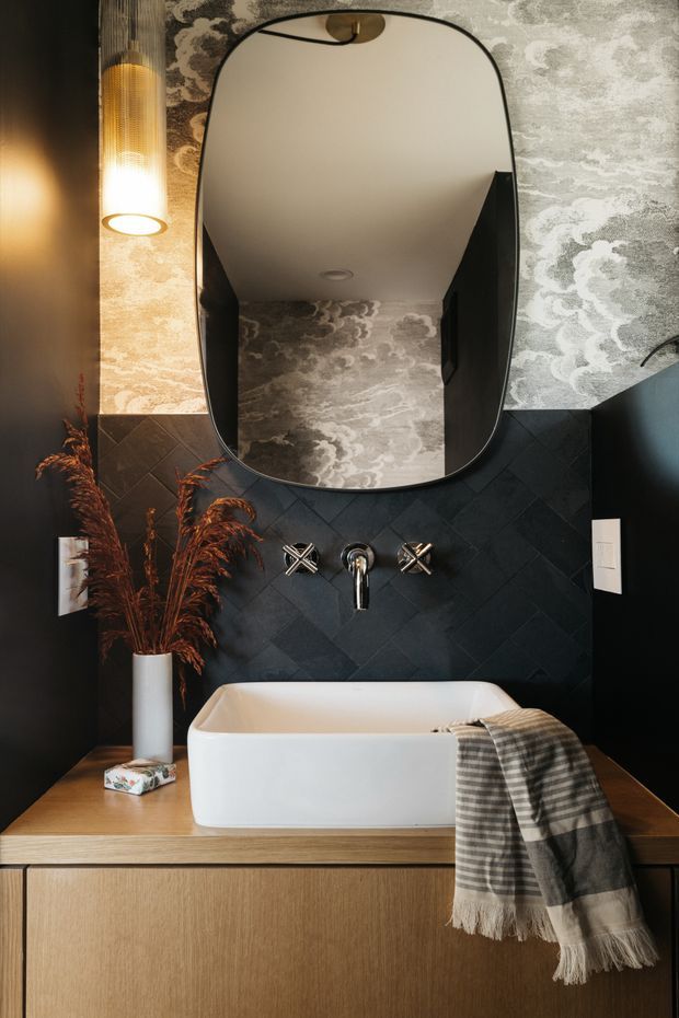 A refined, moody bathroom vanity scene featuring a freestanding white vessel bathtub set into a timber-look vanity cabinet and benchtop. The dark charcoal wall is clad in diagonally arranged tile, while patterned grey-and-cream wallpaper frames a large organic-shaped black-edged mirror. A warm vertical pendant light, wall-mounted tapware, a white vase with dried foliage, and a draped checked textile complete the composition.