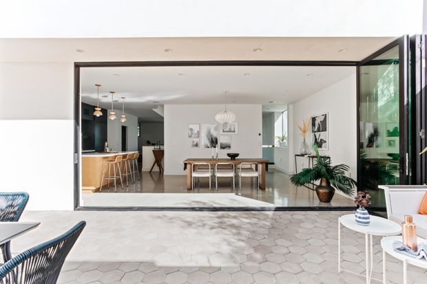 A bright contemporary indoor-outdoor living and dining area viewed from a tiled terrace through fully opened black-framed glazed doors. Inside, a timber kitchen with island and bar stools sits to the left, while a dining table with pendant lighting occupies the centre. The interior uses white walls, pale flooring and restrained decor, with tropical plants and sculptural artwork adding warmth. The foreground terrace has light grey hexagonal tiles, outdoor lounge furniture and small side tables.