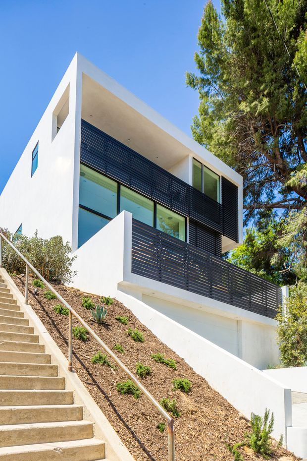 Exterior photograph of a modern multi-storey white house on a steeply sloping site. The angular flat-roofed facade features extensive glazing, deep recessed openings, and dark horizontal louvred screening and balustrades. A long concrete stairway with metal handrails climbs alongside a landscaped bank, while mature trees frame the building.
