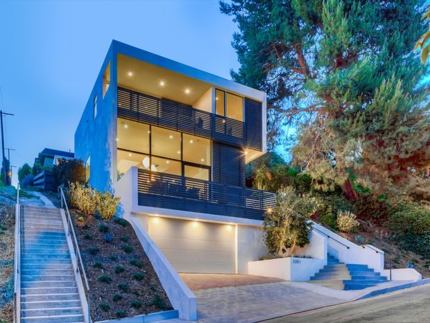 Twilight exterior view of a narrow, multi-storey contemporary residence on a sloped urban site. The home features extensive floor-to-ceiling glazing, dark horizontal balcony and privacy screens, a projecting flat roof, rendered light-coloured walls, an integrated double garage, and stepped pedestrian access with landscaped garden beds and mature trees.