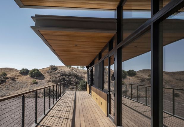 Exterior view of a contemporary building’s covered timber deck or balcony, with expansive dark-framed glazing reflecting the surrounding dry, hilly landscape. The projecting roof has a warm timber soffit, and slim black metal balustrades line the deck. The composition highlights the building envelope, outdoor circulation space, and connection to the rugged natural setting.