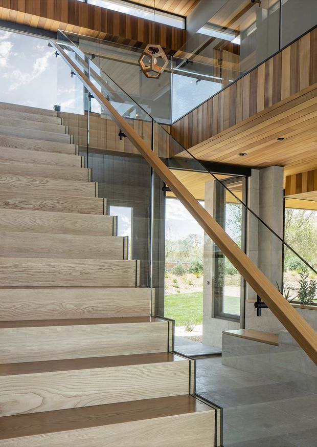 Contemporary interior stairway with broad pale timber-clad treads, a sculptural timber handrail, frameless glass balustrades, dark metal detailing, and extensive floor-to-ceiling glazing. Warm timber wall and ceiling panelling contrasts with exposed grey concrete columns and a polished grey floor. The stair connects multiple levels and is illuminated by linear skylights and large windows overlooking landscaped grounds.
