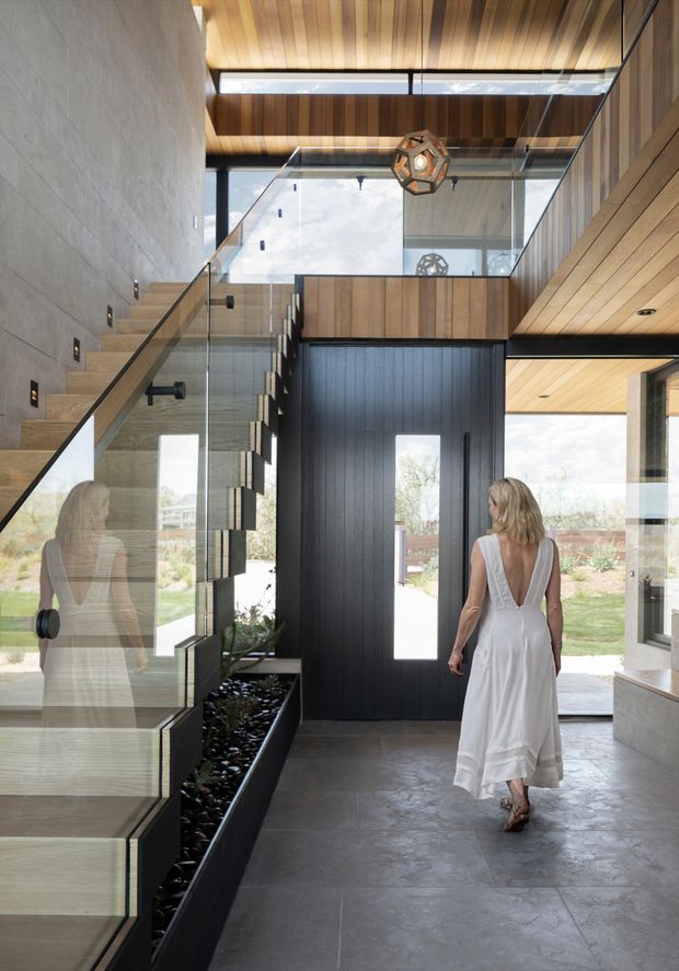 A contemporary residential entryway viewed from inside, featuring a sculptural floating timber staircase with clear glass balustrades, a full-height dark vertical-panel door, expansive glazing, and a warm timber-lined ceiling. A narrow indoor planter with dark stones runs beneath the staircase. The space has a dramatic double-height volume and strong indoor-outdoor connection, with concrete-look flooring and a geometric pendant light.