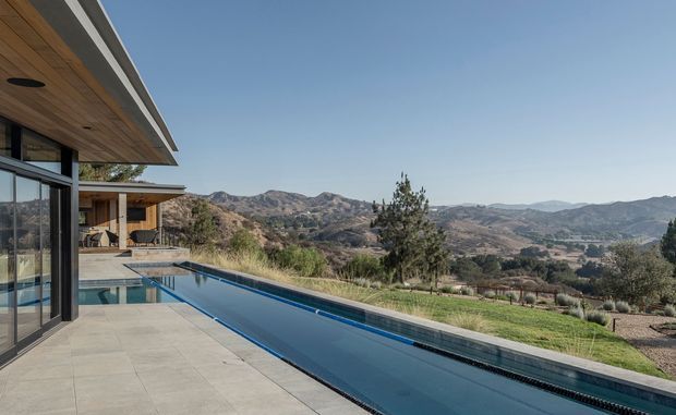 Exterior architectural view of a contemporary house beside a long narrow infinity-edge swimming pool. The house features broad roof overhangs, warm timber soffits, dark slender window and door frames, extensive glazing, and pale stone or concrete terrace paving. The pool overlooks a dry, rolling mountainous landscape under a clear sky.