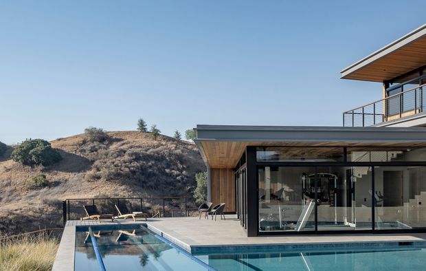 Exterior view of a contemporary multi-level home on a dry, sloping site, with extensive black-framed glazing, timber soffits, flat roof forms, and a balcony overlooking a long rectangular swimming pool and paved pool terrace. A gym or fitness room is visible through the glazing. The setting includes a rugged, sparsely vegetated hillside.