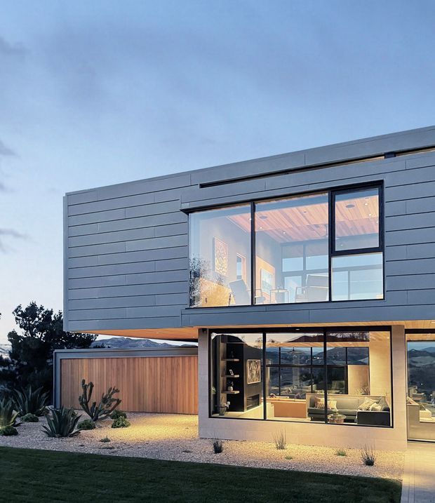 Contemporary two-storey house exterior at dusk, featuring a cantilevered upper volume clad in dark horizontal panels, extensive black-framed glazing, warm timber soffits, and a timber-clad garage or boundary structure. The illuminated lower-level living area opens toward a landscaped garden with gravel, sculptural planting, and lawn; mountains are visible in the distant background through the glazing.