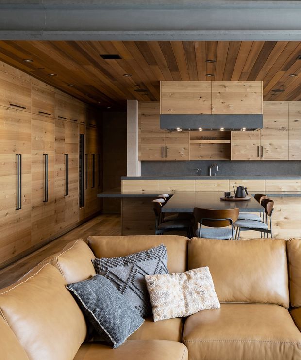 A warm open-plan living and kitchen space dominated by extensive light timber cabinetry and a timber-lined ceiling. The foreground contains a large caramel leather sectional sofa with patterned cushions, while the background features a compact dining table with upholstered chairs, a dark benchtop, integrated appliances, and a double-bowl sink. Recessed downlights are set into the timber ceiling, and tall built-in storage runs along the left wall.