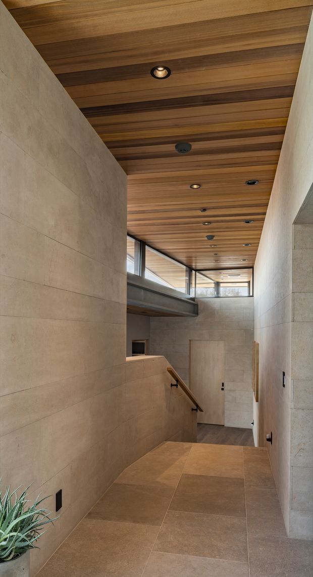 A tall, narrow modern interior hallway and stair landing with warm wood slat ceiling, recessed downlights, large beige stone-look wall panels, and large-format beige floor tiles. A stair descends toward a timber-finished lower level, with high clerestory glazing bringing in natural light. The space has a refined, warm contemporary architectural character.