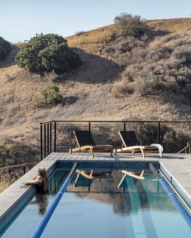 An outdoor lap swimming pool set into a dry, rolling hillside landscape. The pool has dark blue water, lane dividers, pale paved coping, and a safety fence along the rear edge. Two reclining sun loungers sit on the deck, while scrub-covered slopes and mature shrubs form the backdrop.
