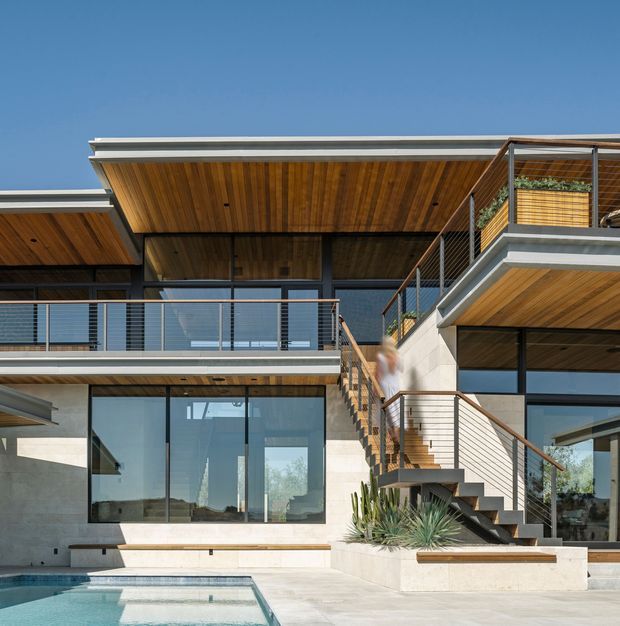 Contemporary multi-level residential facade with expansive dark-framed glazing, layered flat roof planes, warm timber soffits, concrete and pale masonry walls, and multiple terraces with slender metal balustrades. An exterior stair connects the levels, while a swimming pool and drought-tolerant planting occupy the foreground courtyard.
